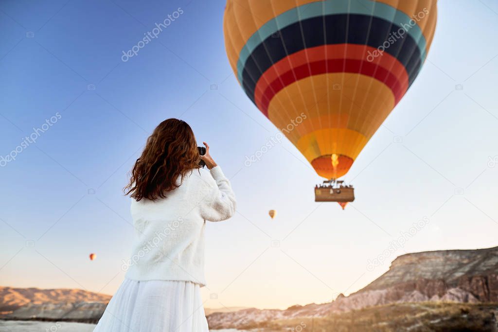 A tourist photographer girl wearing white sweater on a mountain top enjoying wonderful view and taking a picture of balloons in Cappadocia. Travel photographer in Turkey