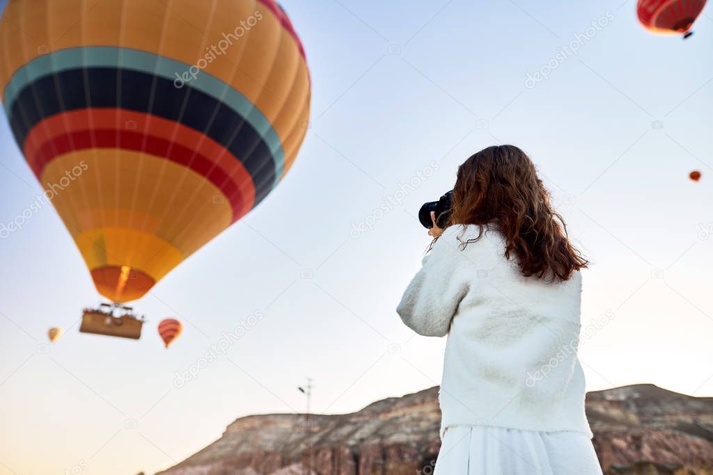 A tourist photographer girl wearing white sweater on a mountain top enjoying wonderful view and taking a pictures of balloons in Cappadocia. Travel photographer in Turkey