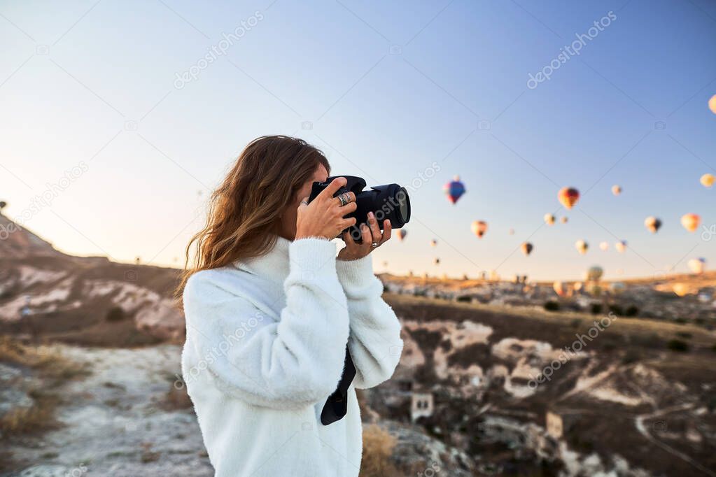 A tourist photographer girl wearing white sweater on a mountain top enjoying wonderful view and taking a pictures of balloons in Cappadocia. Travel photographer in Turkey