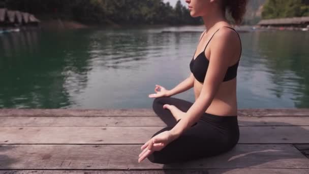 Женщина сидит у озера и практикует йогу — стоковое видео