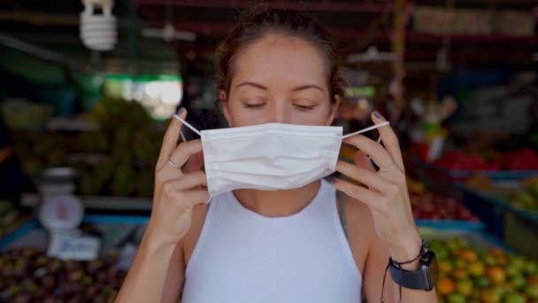 Une jeune femme porte un masque de protection, se tient au marché et regarde la caméra — Video