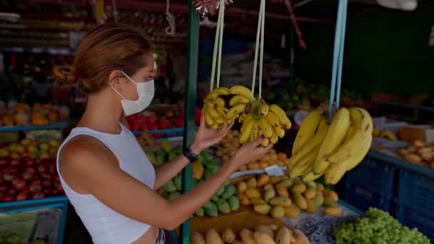 Una joven con una máscara protectora compra fruta en el mercado — Vídeo de stock
