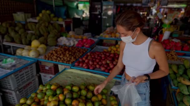 一位戴防护面具的年轻女子在市场上买水果 — 图库视频影像