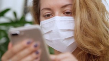 Beyaz koruyucu maske takan genç bir kadın akıllı telefondan haber okuyor.