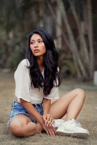 Vackra asiatiska kvinna säten på gräs, bär jeans shorts, leende njuta av helger — Stockfoto