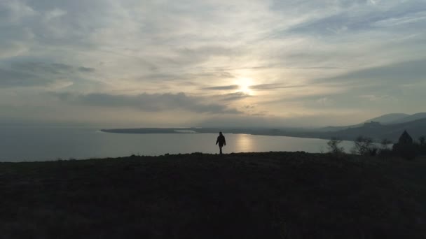 Один турист идет по холму к озеру на закате — стоковое видео