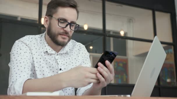 Мужчина улыбается и использует телефон для чата в офисе — стоковое видео