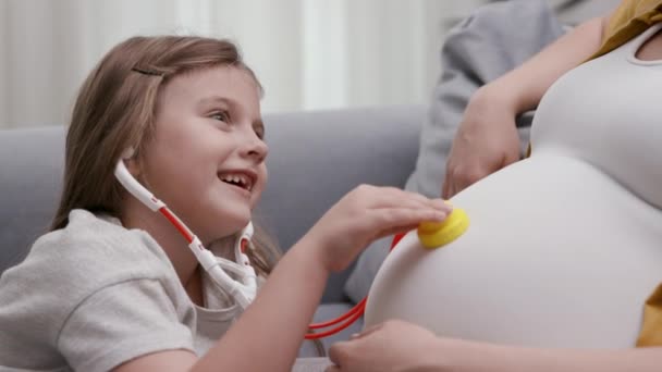 Γλυκό κοριτσάκι χαϊδεύει παιχνιδιάρικα τη μαμά έγκυο κοιλιά με το παιχνίδι ιατρική φροντίδα — Αρχείο Βίντεο