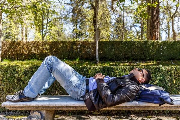 A man in leather jacket and jeans is resting on a bench in a par