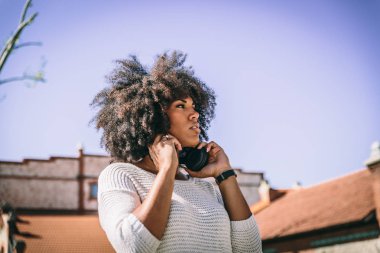 Beautiful afro brunette girl puts on black headphones while walking through a park