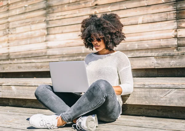 Afro stil kızı dizüstü bilgisayarıyla çalışırken bacak bacak üstüne atmış bir şekilde parkta oturuyor.