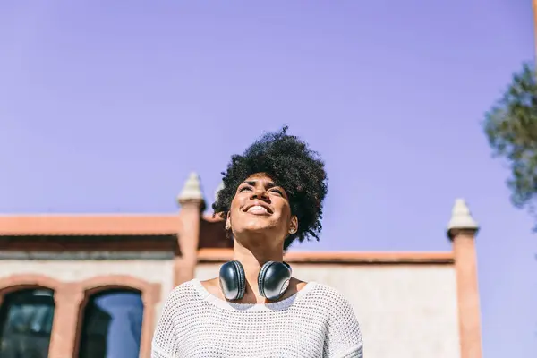 Beautiful dark skinned woman with big black headphones around her neck, smiles looking up while walking through — Stock Photo, Image