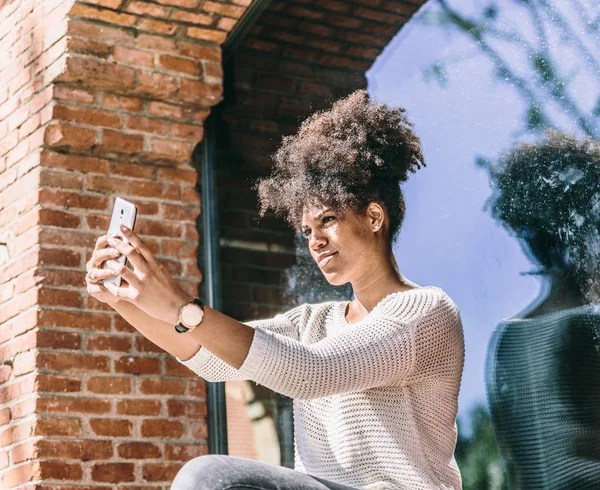 Beautiful young afro haired girl takes a selfie with a smile, next to a large window