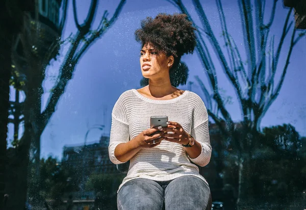 Pretty brunette girl with afro hair sitting by a window with a smartphone in her hands — Stock Photo, Image