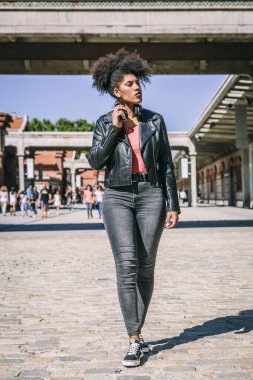 Saçlı, siyah elbiseli ve kulaklıklı bir kız, bir caddenin ortasında huzur içinde yürüyor.