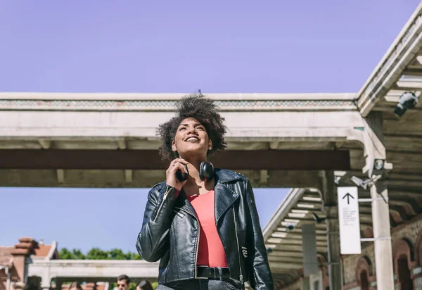 Woman with dark skin, afro hair with casual clothes and headphones around her neck, smiles while looking straight ahead when walking — Stock Photo, Image