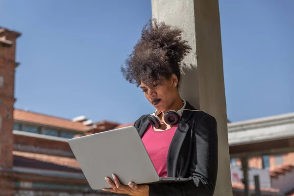 Attractive woman with black jacket, headphones around neck and afro hair, works with her laptop resting on a column in a street