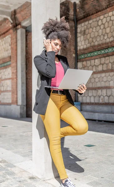 Attractive brunette woman with black jacket leaning on a column in the street, checks something on her laptop while listening to music