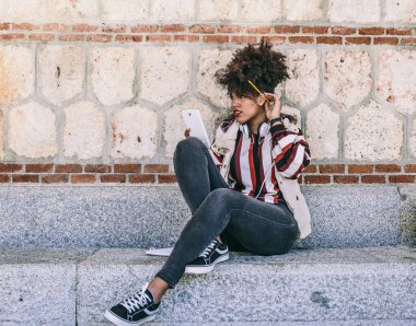 Afro saçlı esmer bir öğrenci elinde tablet tutarken bir şey düşünüyor ve taş bir bankta oturuyor.