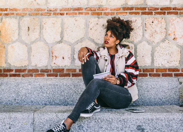 Beautiful brunette girl with afro hair is sitting on a stone bench, thinking something to write in her notebook, on an autumn day — Stock Photo, Image