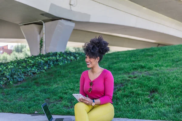 Beautiful and smiling entrepreneur girl with afro hair, is sitting in a quiet park working relaxed with her tablet and laptop.