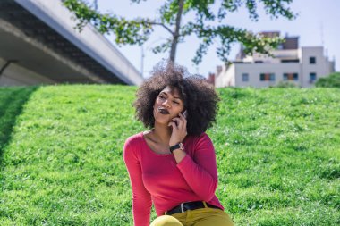Afro saçlı bir kadın sessiz bir bölgede akıllı telefonuyla konuşurken yaramaz bir yüz takınır. 