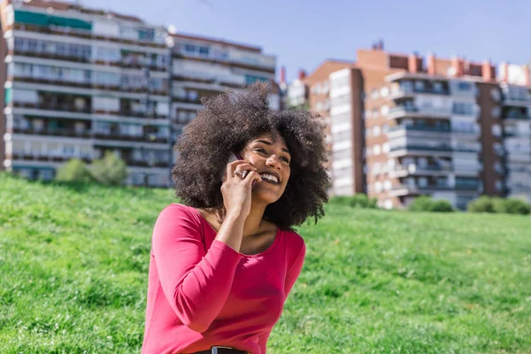 Cute afro woman speaks happily with her mobile phone, sitting on the grass of a large park — Stock Photo, Image