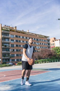 Kollarında basketbol topuyla atletik bir adam şehir sahasında basketbol potasının yanında.
