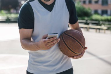 Bir elinde basketbol topu, diğer elinde akıllı telefonu olan bir sporcunun yakın çekimi.