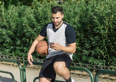 Topunun yanındaki basketbolcu dışarıda otururken akıllı telefonunu kontrol ediyor.