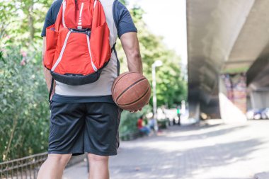 Topunu sırtında taşıyan bir erkek basketbolcunun ve sırtında parkta yürüyen büyük kırmızı bir sırt çantası.