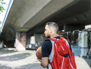 Büyük kırmızı bir sırt çantası ve yürürken basketbol topu taşıyan güçlü bir basketbolcunun arkası.