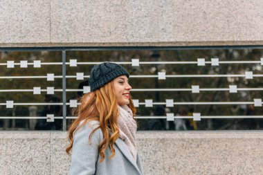 Şapkalı, atkılı ve ceketli genç sarışın kız camlı gri duvarın yanında gülümsüyor.