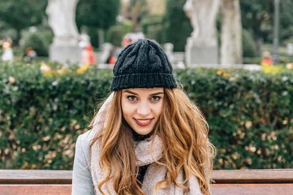 Belle fille blonde européenne souriante avec un chapeau en laine assise sur le banc d'un parc de la ville — Photo