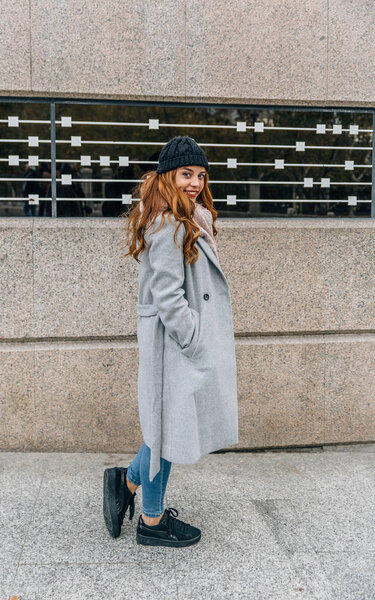 Urban style blonde girl dressed in gray coat and woolen hat walks smiling on a street