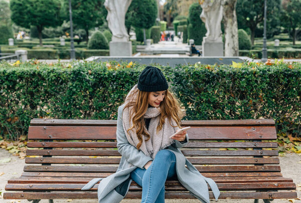 Caucasian blonde girl with coat, scarf and hat, checks her smartphone while sitting on a park bench