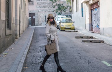 Caucasian elegant woman wearing white coat and hat crosses a city street