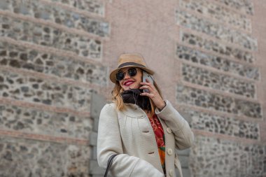 Very attractive blonde woman smiling with coat and black glasses speaks on her mobile phone by a city