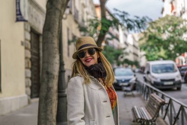 Attractive colorful woman with coat, hat, scarf and black glasses smiles happily on a street
