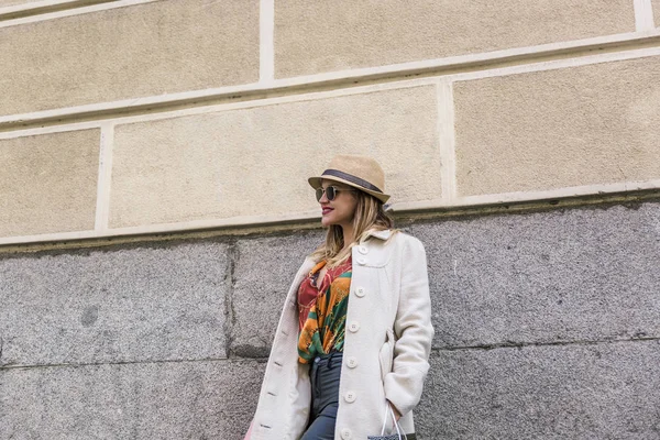 Cute and attractive blonde woman with white coat and colorful clothes leaning on a wall