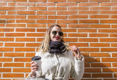 Beyaz ceketli, tuğladan duvara yaslanmış siyah atkısı olan kahve fincanı taşıyan beyaz bir kadın.