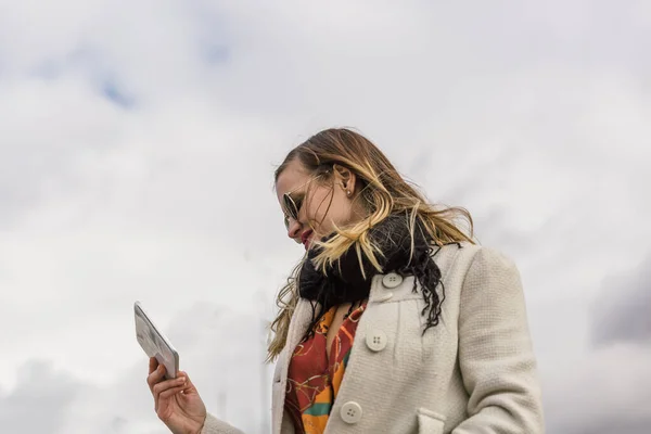 Fille blonde caucasienne avec manteau blanc et des vêtements colorés regarde sa tablette dans la rue — Photo