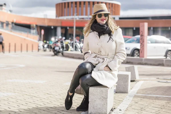Mujer rubia caucásica muy atractiva con sombrero y abrigo blanco sonríe sentada junto a una gran estación — Foto de Stock