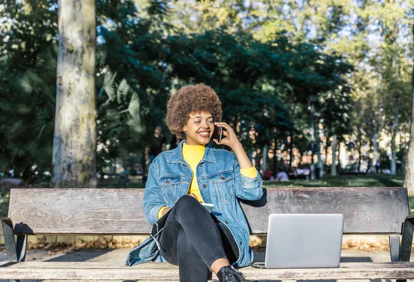 Beautiful smiling girl with afro hair speaks on her mobile phone sitting on the bench of a large park — Stock Photo, Image
