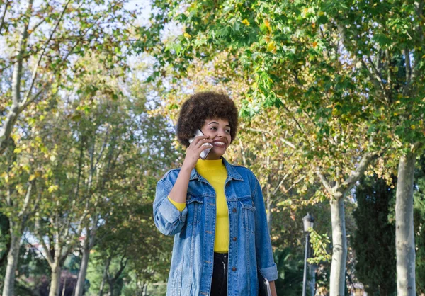 Young afro girl with perfect smile and white teeth smiles while talking with her smartphone while walking — Stock Photo, Image