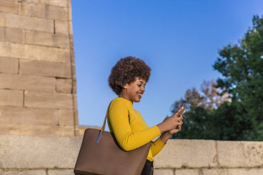 Afrika saçlı modern melez kız, güneşli bir günde cep telefonuyla mesajlaşırken gülümsüyor.