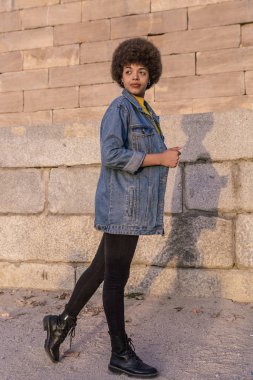 Afro saçlı şık esmer kadın mavi kot gömlek ve siyah pantolonla sokakta yürüyor.