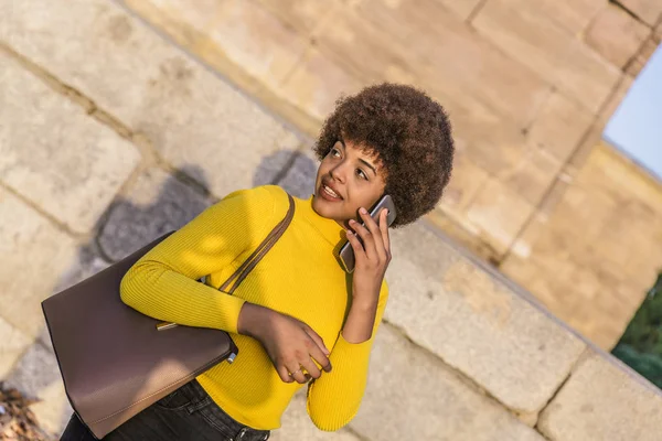 Attractive mixed race business woman with afro hair and yellow shirt talking on her smartphone in the middle of a city — Stock Photo, Image