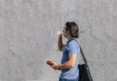 Uzun siyah saçlı genç adam su içerken diğer elinde kırmızı bir termos vardı, gri bir duvarın yanından geçiyordu.