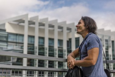 Gülümseyen ve mutlu beyaz bir adam bir bahar gününde metal bir korkuluğa yaslanıyor ve şehrin dışında geziyor.